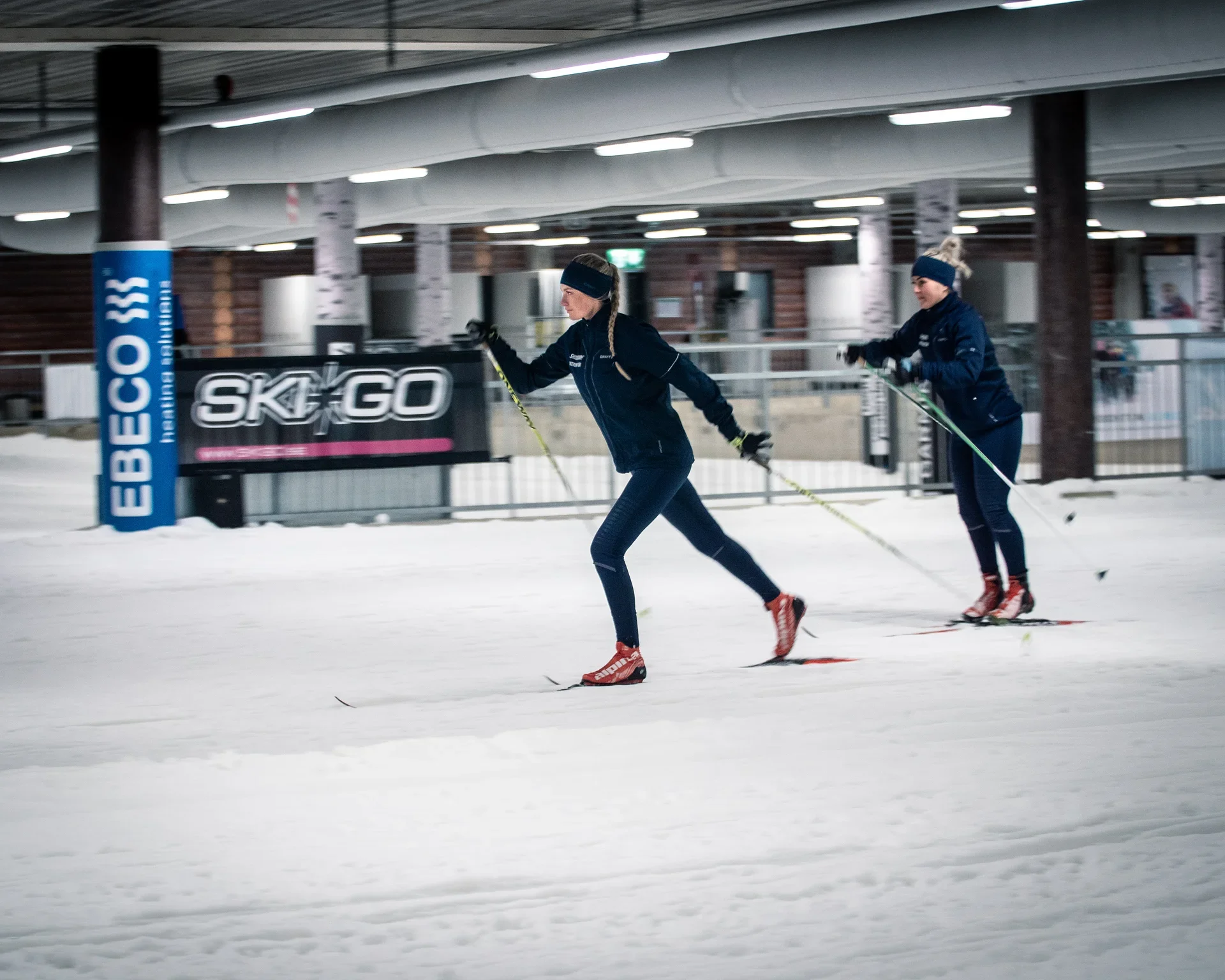 Inomhus skidanläggning med snöspår, skidor och moderna träkonstruktioner.